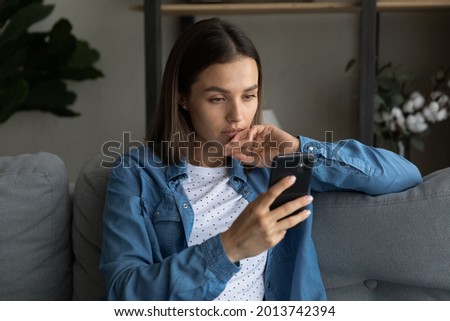 Serious thoughtful 25s woman sit on sofa at home holding smartphone device read sms feels upset, got unpleasant news, thinks over received notification internet information, learn new software concept Royalty-Free Stock Photo #2013742394