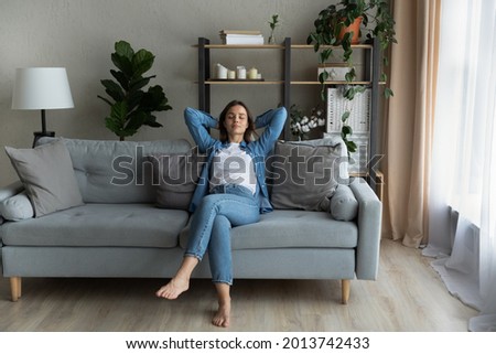 Serene young beautiful woman relaxing on comfy sofa with hands behind head, breath fresh conditioned air inside of modern light living room. Modern house, stress-free weekend, contemplation concept Royalty-Free Stock Photo #2013742433