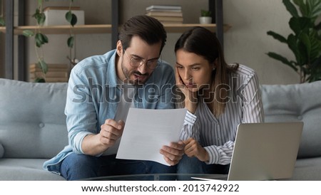 Serious pensive millennial 35s couple sit on sofa at home hold letter reading papers documents, learn details of contract, discuss agreement terms conditions, considering mortgage loan offer concept Royalty-Free Stock Photo #2013742922