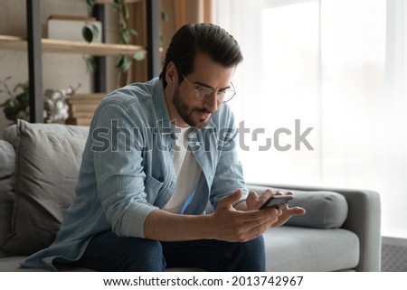 Serious millennial man in glasses sit on sofa holding smartphone check online calendar, sending message on messenger answers to friend, using business mobile app. Modern tech usage, leisure concept