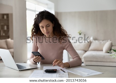 Young Hispanic woman sit at desk at home manage household finances paying bills calculating on machine cellphone. Millennial Latin female care of budget use smartphone for payment. Finance concept. Royalty-Free Stock Photo #2013743036