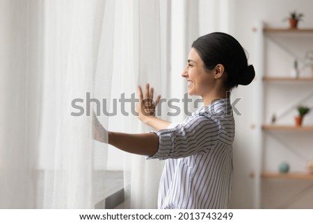 Side view smiling dreamy Indian woman opening curtains in early morning, looking out window in distance, thinking and dreaming, happy beautiful young female feeling positive, starting new day Royalty-Free Stock Photo #2013743249