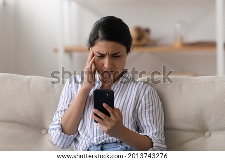 Unhappy Indian millennial woman looking at phone screen, sitting on couch, reading bad news in message, upset young female holding smartphone, having problem with broken or discharged mobile device Royalty-Free Stock Photo #2013743276