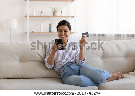 Excited Indian woman paying online by credit card for goods, using smartphone, sitting on couch at home, happy young female satisfied customer browsing internet banking service on device, shopping Royalty-Free Stock Photo #2013743294