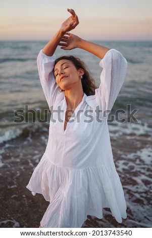 Young happy woman in a white fluttering dress walks along seashore. The girl looks at the magical sunrise. Summer time. Travel, weekend, relax and lifestyle concept.