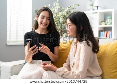 Two pretty best friends of Asian Korean beauty spend an afternoon on the living room couch, talking, the woman shows vacation photos on her phone, they look together, smile, comment