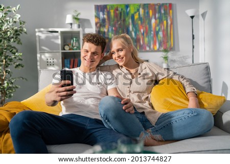 A couple of attractive teenagers in love are spending their free time after school together at home on couch, man with curly hair is holding phone, taking a picture with a smiling beautiful blonde