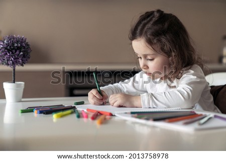 Little happy girl sitting at a table at home in the kitchen and draws a picture with colored pencils and markers