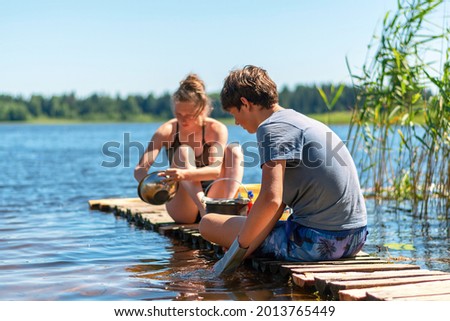 A young girl and a boy are washing dishes on a wooden bridge by the lake. Teenagers on vacation. Summer background. Camping. Royalty-Free Stock Photo #2013765449