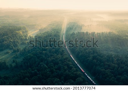 Train in summer morning forest at fog sunrise. Aerial view of moving freight train in forest. Morning mist landscape with train, railroad, foggy trees. Top aerial drone view near railway station. Royalty-Free Stock Photo #2013777074