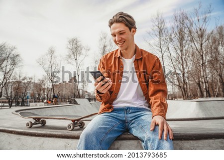 A guy sits on the edge of a ramp in a gray skatepark near a skateboard and looks at the phone, checks messages, chats with friends, browses social networks, publishes photos with his workout 