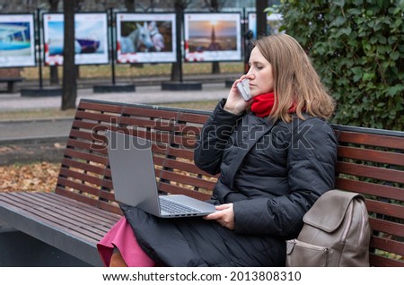 girl works on the computer while sitting on a park bench and speaking by the phone