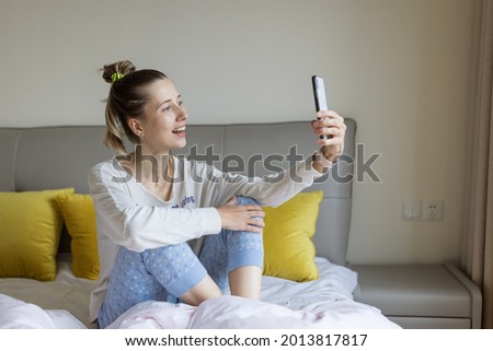 Young caucasian woman using mobile phone and wireless headphones, resting, sitting on bed at home and listening podcast or clubhouse - voice-only social media app, drop-in audio chat