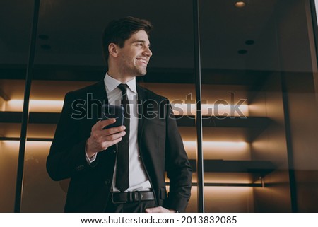 Bottom view fun happy employee agent business corporate lawyer in formal black suit shirt tie use mobile cell phone look aside indoors illuminated apartment wall Achievement career lifestyle concept