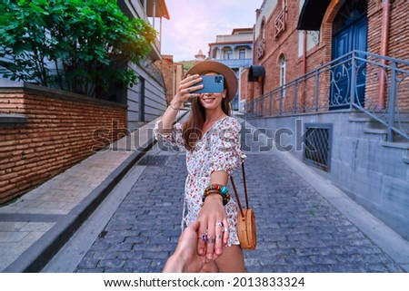 Follow me concept and traveling together. Happy smiling joyful young girl traveler holds the boyfriend's hand and taking photo on a phone camera during vacation weekend time while sightseeing 