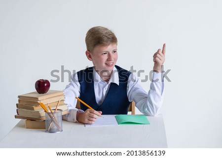 Eureka, a handsome schoolboy in a school uniform, sits at a table next to a stack of books, apples and pencils. Surprised, inspired Raises the index finger up, on a white background