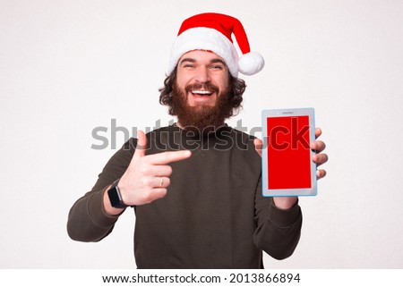 Cheerful smiling bearded hipster man wearing santa claus hat pointing at blank screen on tablet.