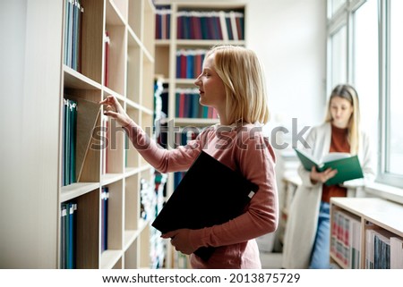 Smiling college student choosing book from bookshelf in a library.