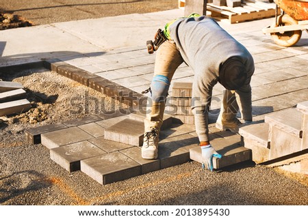 Landscape worker paving driveway interlock patio with stone brick at construction site. Contractor wearing safety protective cloth, gloves and knee pads for heavy installation yard work project. Royalty-Free Stock Photo #2013895430
