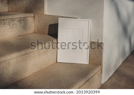 Blank white picture frame leaning against white wall. Outdoor sandstone stairs in sunlight, shadows overlay. Empty poster mockup for art display. Minimal summer design. No people. 