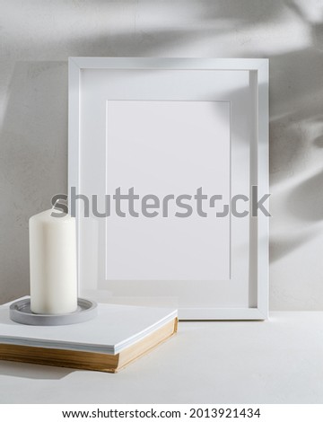 Empty white frame with a passe-partout, a white candle on a concrete plate, and a stack of books in a light interior. Mockup with a place for your design, illuminated by sunlight.