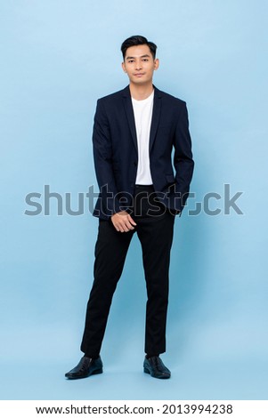 Full length portrait of smiling young handsome Asian man in semi formal suit standing with one hand in pocket on light blue studio background Royalty-Free Stock Photo #2013994238