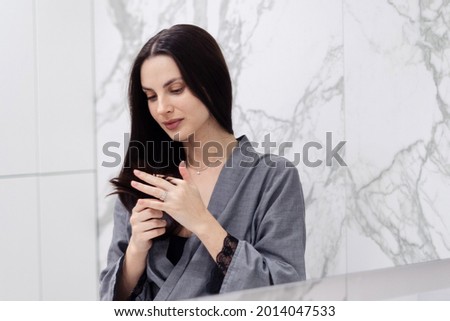 Beautiful brunette young woman looking at her perfect healthy hair in bathroom during selfcare evening routine. Treatment for prevention of split ends and breakage Royalty-Free Stock Photo #2014047533