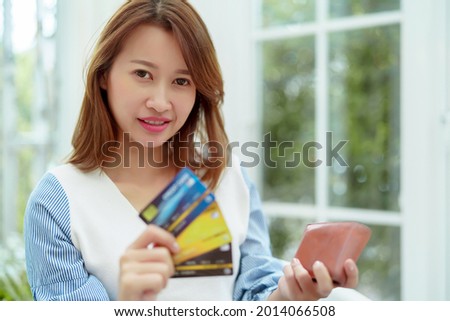 Portrait of a beautiful Asian woman in a white shirt sitting in front of a computer, taking out her credit card from her purse, preparing for online shopping with a happy smile on her face.