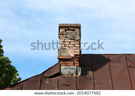 Red building bricks dilapidated narrow chimney with cracked broken facade and visible metal protection sheet on edge of abandoned suburban family house roof covered with rusted metal roof tiles Royalty-Free Stock Photo #2014077482