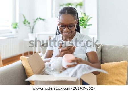 Young satisfied happy african girl woman lady shopaholic customer sit on sofa unpack parcel delivery box, online shopping shipment concept. taking photos of product to post on social media