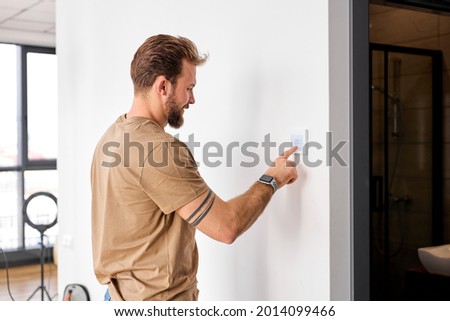 Handsome man standing near wall-mounted device adjusting degrees in living room set comfortable temperature using thermostat home heating system. Owner of modern smart house, energy saving concept Royalty-Free Stock Photo #2014099466