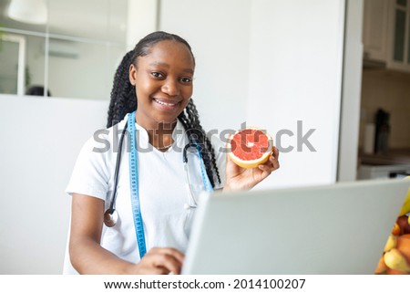 Portrait of young smiling female nutritionist in the consultation room. Nutritionist desk with healthy fruit, juice and measuring tape. Dietitian working on diet plan.