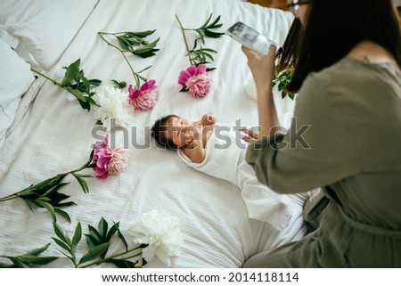 Mother blogger taking photo portrait of her cute newborn baby girl sleeping surround with flowers peonies at cozy white bedroom at home.