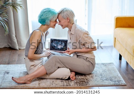 Pregnant lesbian couple with sonogram on floor in living room, side view portrait of two short-haired ladies in casual wear feeling love, happy to be future parents moms. copy space. lgbt, pregnancy Royalty-Free Stock Photo #2014131245