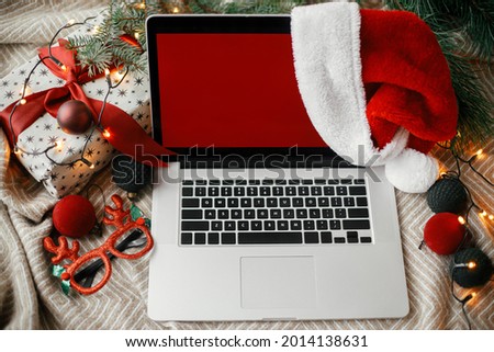 Laptop with red empty screen in santa hat on cozy bed with stylish christmas ornaments, present and festive lights. Christmas sales and shopping online. Space for text. View above. Freelance