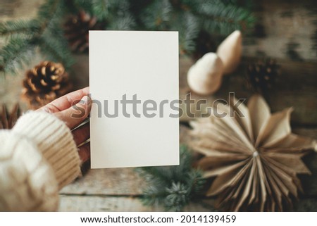 Christmas card mock up. Hand holding empty greeting card on background of christmas paper stars, wooden tree, pine branches and cones on rustic wood.  Space for text. Seasons greetings template