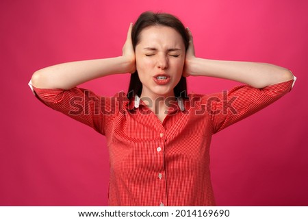 Beautiful young woman gestures she doesn't want to hear and holds hands on her ears against pink background