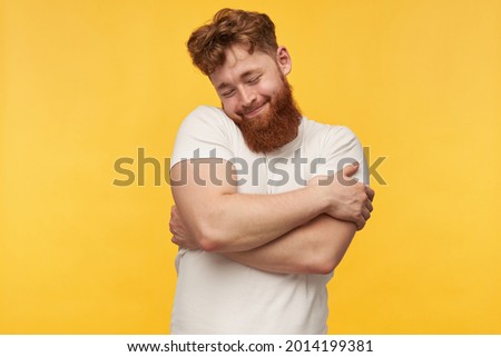 indoor portrait of young bearded guy, wears blank t-shirt, keeps his eyes closed, hug himself and smiling. isolated over yellow background
