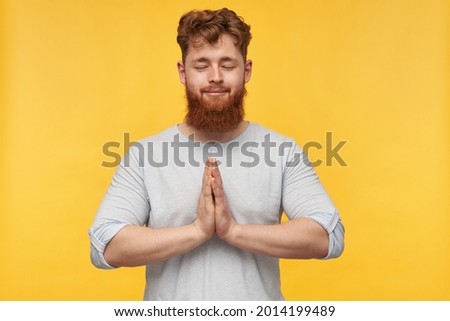 young bearded guy keep calm, keeps his palm together, eyes closed and smiles. meditation after long day. isolated over yellow background.