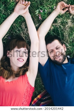 Couple makes a selfie while lying on the grass in flowers. Rest, lovers on a flower meadow. Bearded man and a girl in a red dress take pictures of themselves in the field. Husband and wife on vacation