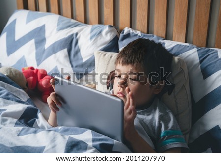 Kid reading bed time stories on tablet before sleep, Happy boy sitting in bed playing games on digital pad, Child relaxing at home in his bed room on weekend.Children with technology concept