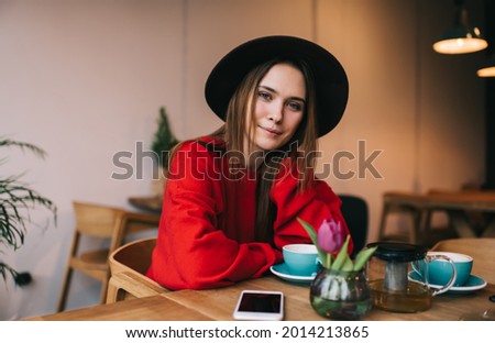 Cheerful female with brown hair in red sweater and black hat sitting at table with cups of tea and looking at camera in cozy cafe