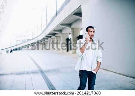 Thoughtful young African American male in casual clothes having conversation on smartphone while walking on street near white bridge in daylight