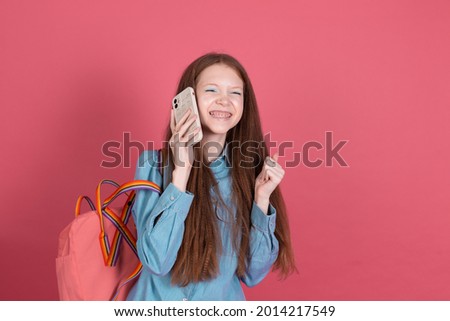 Little kid girl 13 years old in blue denim jacket isolated on red background schoolgirl with backpack with mobile phone happy winner gesture