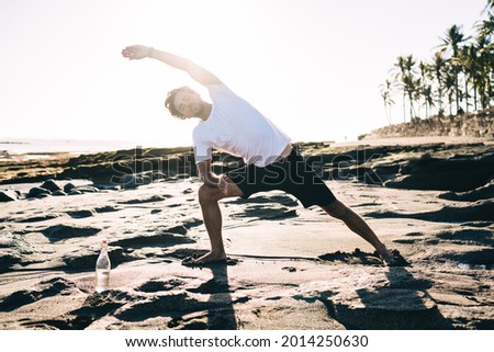 Full length focused barefoot male in sportswear practicing yoga on sandy shore doing Extended Side Angle pose against sea water and palms in back lit