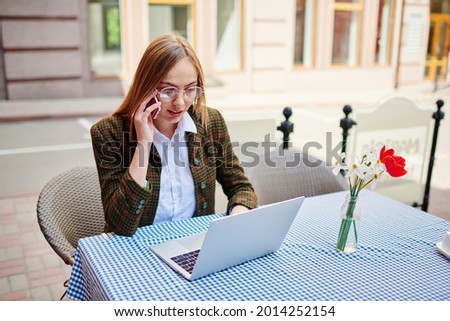 Serious female remote worker in casual clothes sitting at table on street typing on laptop and having conversation on mobile phone