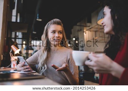 Young woman with blonde long hair talking and actively gesticulating while female friend drinking coffee and listening with bright smile Royalty-Free Stock Photo #2014253426
