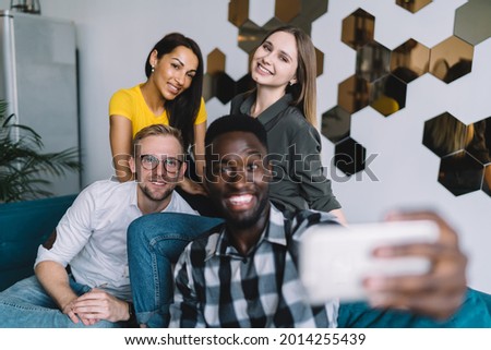 Positive group of diverse friends in casual wear sitting on sofa in modern place and taking selfie on mobile phone
