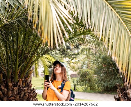 young smiling woman tourist in cap yellow t-shirt with backpack taking photo on smartphone of tropical plant palm tree cycas in park travel using modern technology selective focus