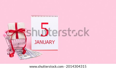 5th day of january. A gift box in a shopping trolley, dollars and a calendar with the date of 5 january on a pink background. Winter month, day of the year concept.
